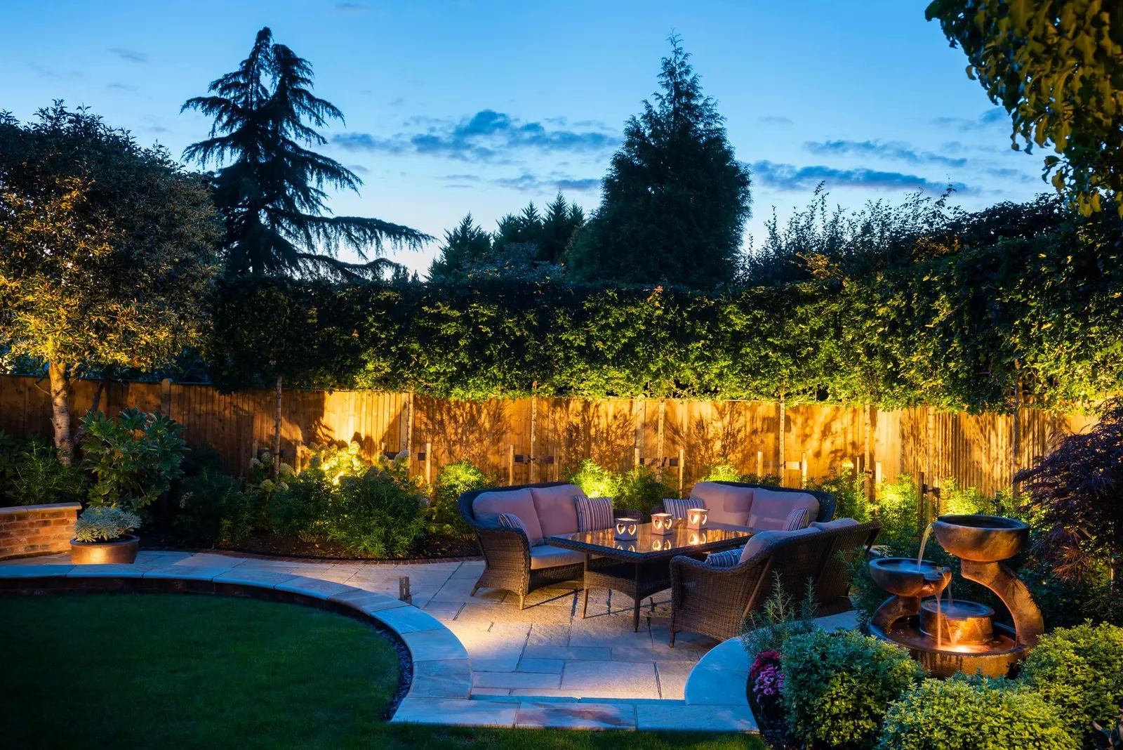 A curved sandstone patio at dusk with a rattan sofa set, warmly uplit fence and hedge behind, and a copper cascading water bowl on the right