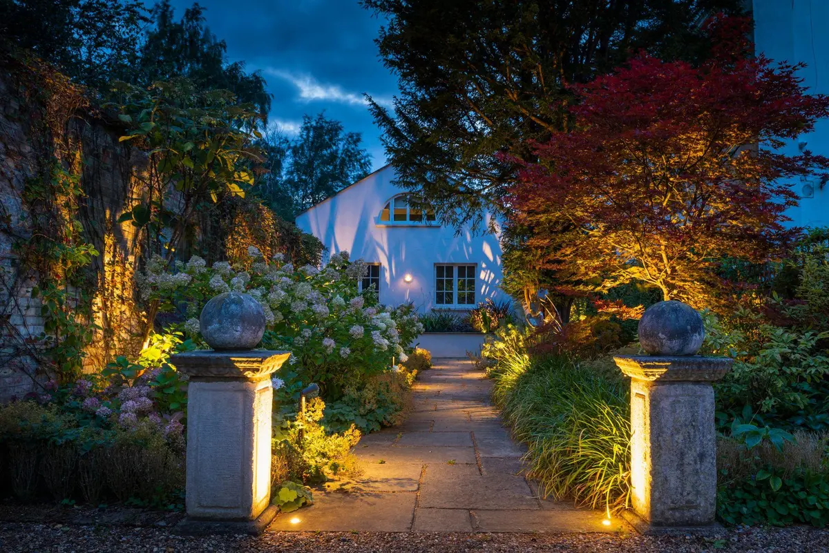 A stone path between two pillars topped with carved orbs at dusk, hydrangeas warmly uplit and a red Japanese maple glowing on the right