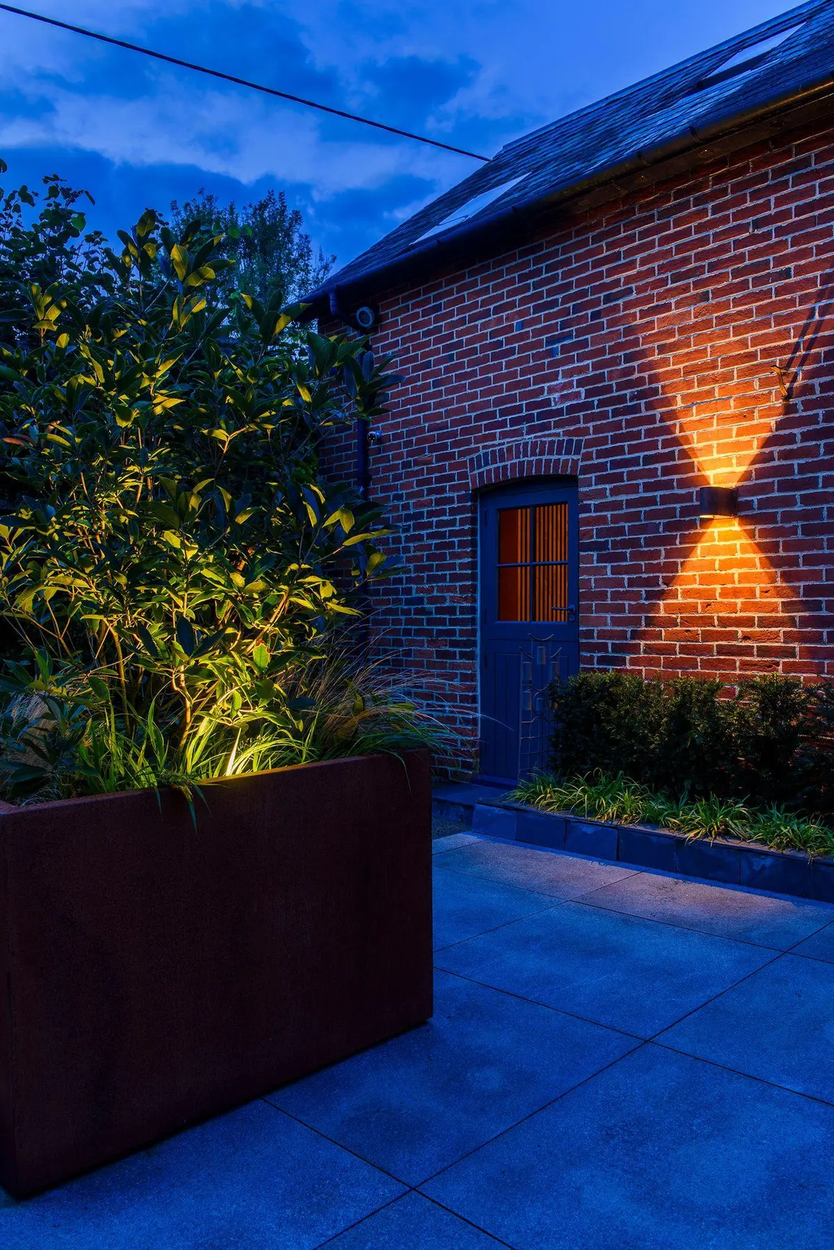 A red brick outbuilding at dusk with an up-and-down wall light casting an hourglass onto the brickwork, and a corten planter with a warmly uplit shrub on the left