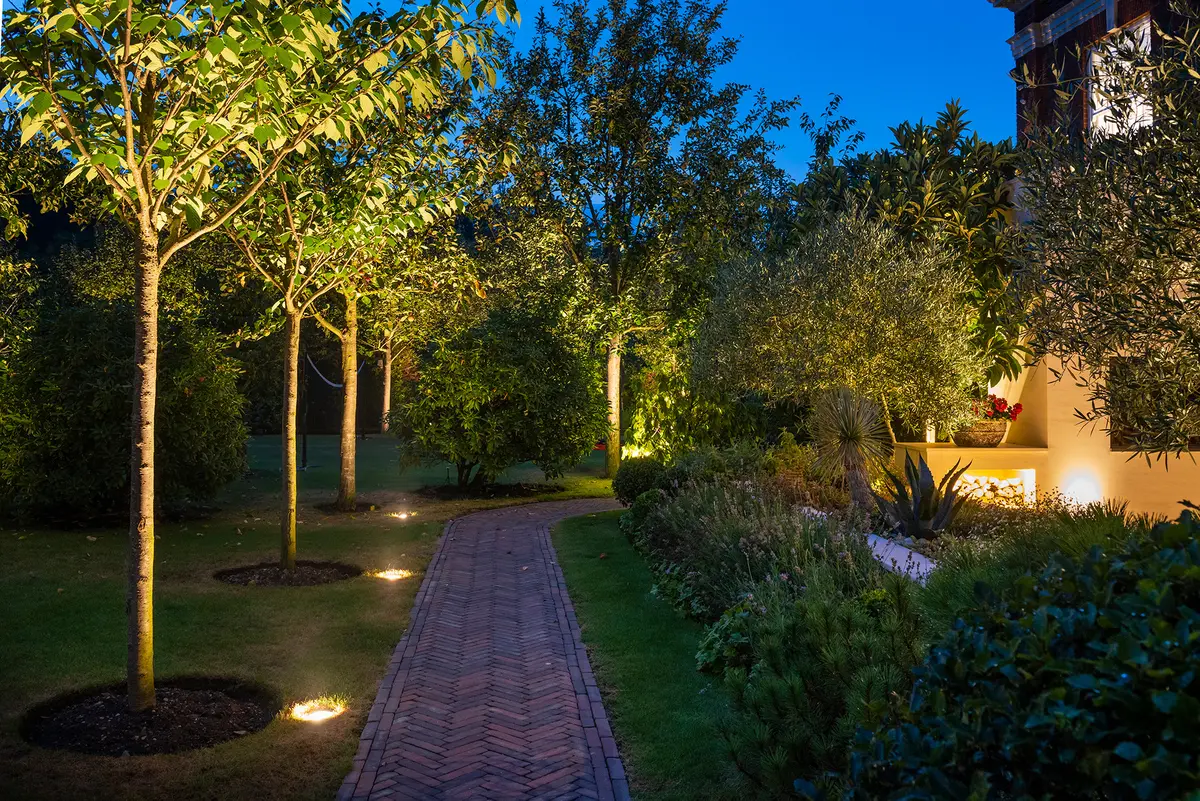 A herringbone brick path winding through a garden with uplit ornamental trees and a lit raised border