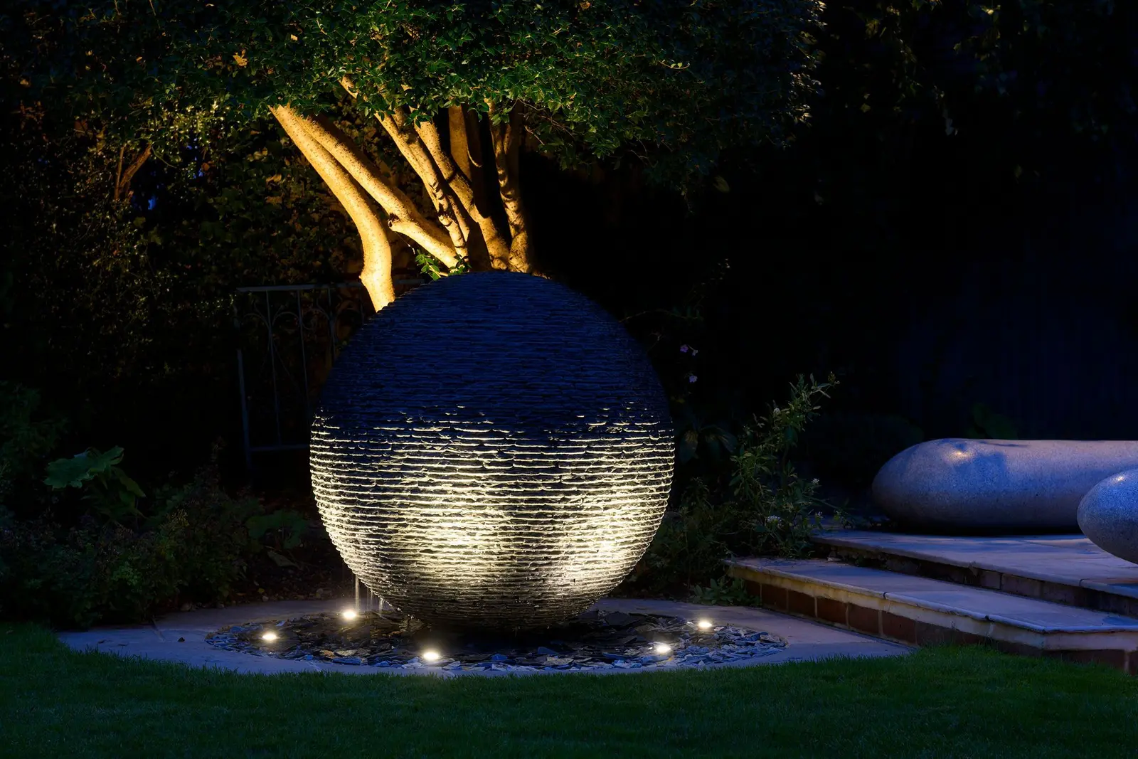 Slate sphere water feature glowing beside ground-level path lights after sunset