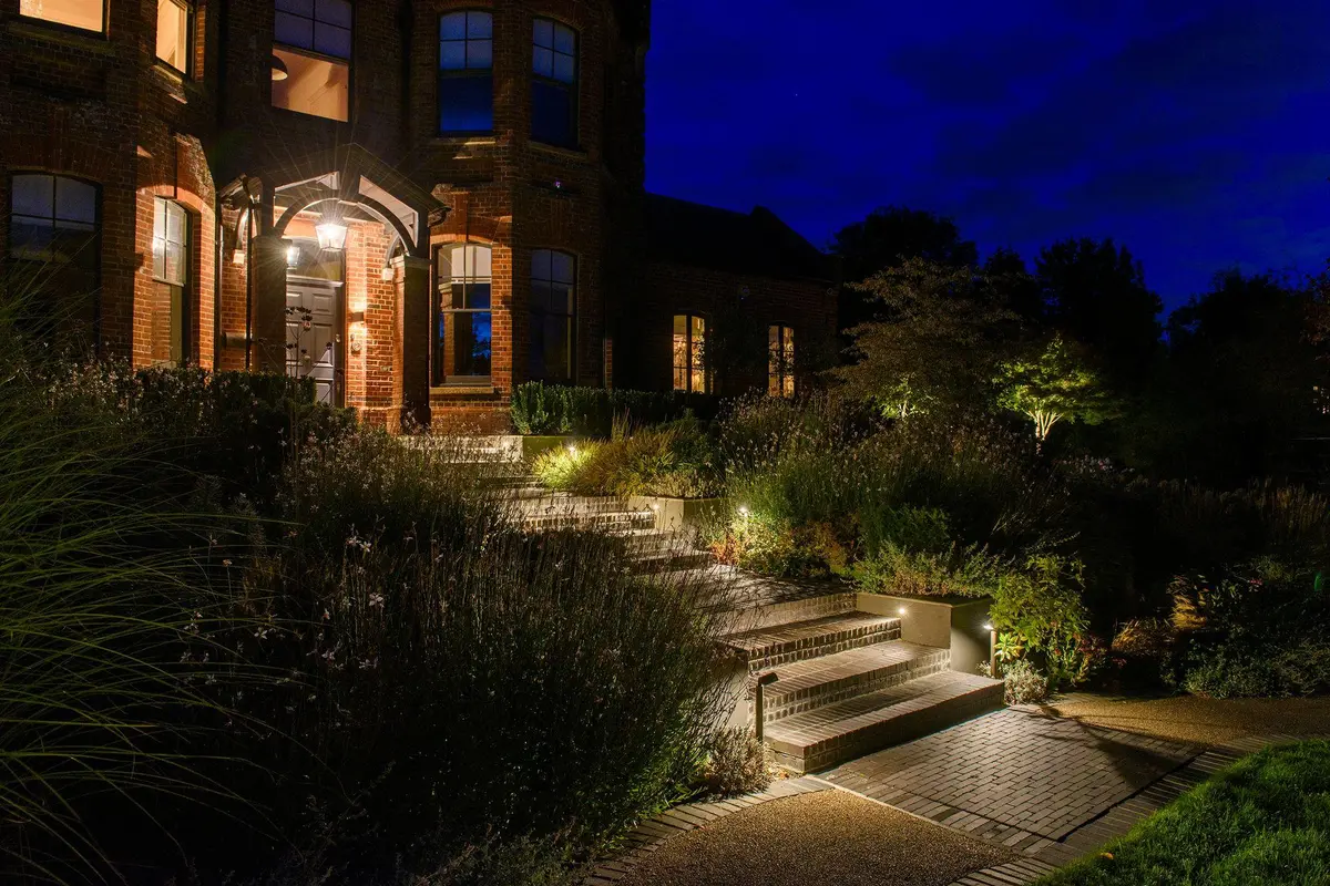 Brick steps rising through planted borders to the front door of a Victorian brick house at dusk, with recessed step lights and warm uplights in the planting