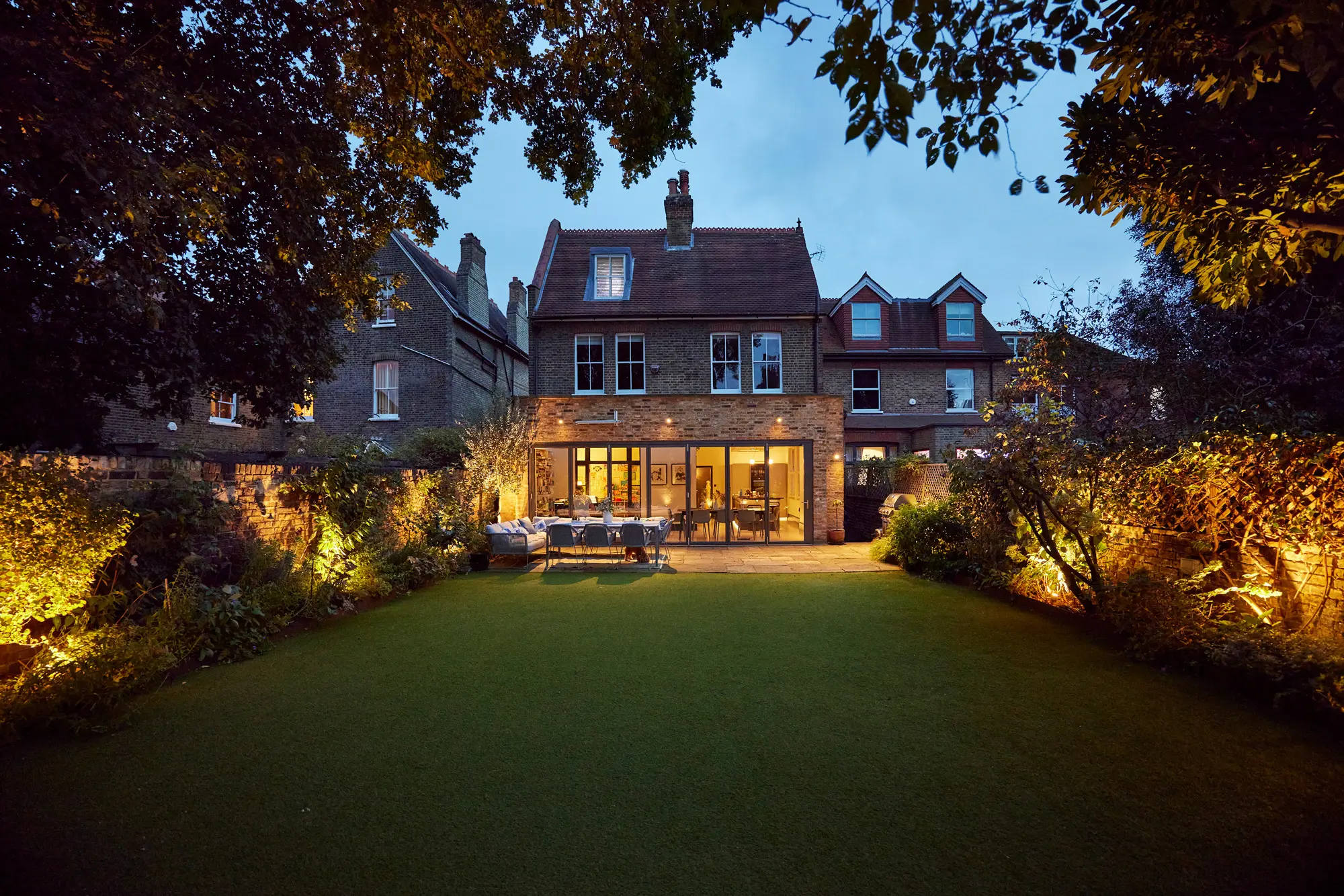 The rear of a Victorian brick home at dusk, with a lit patio, dining set and warmly uplit border planting