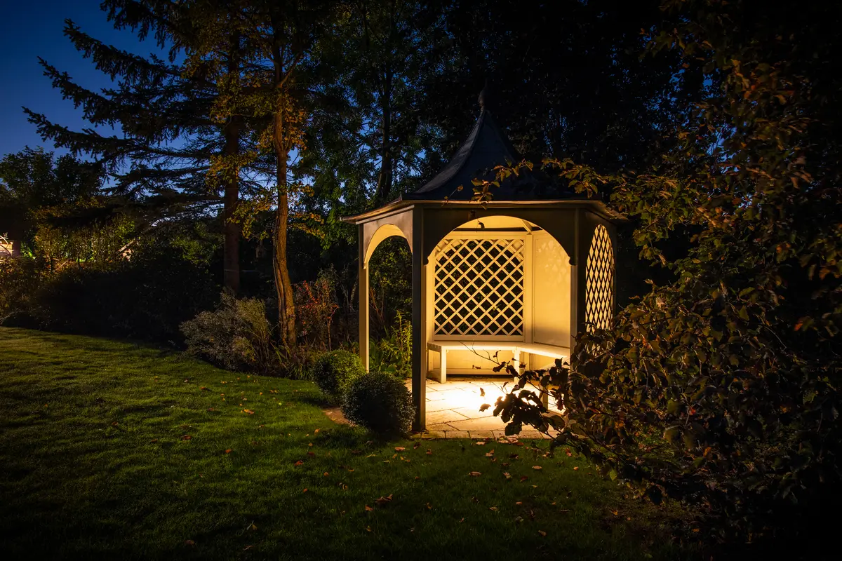 A hexagonal timber gazebo glowing warmly from within, set on a lawn beside mature trees at dusk